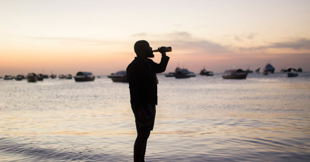 A man enjoys a drink by the ocean at sunset, silhouetted against the sea and sky.