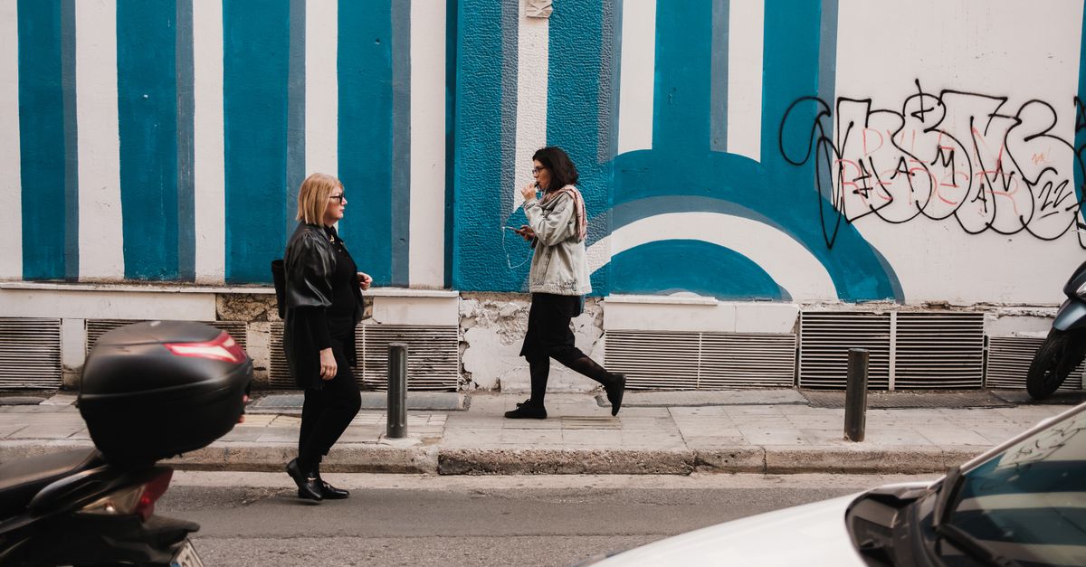 Two women walk past a vibrant blue mural and graffiti on an urban street, surrounded by vehicles.