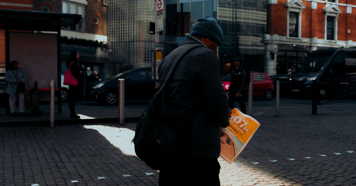 Man in city street scene holding a newspaper with urban buildings in background.