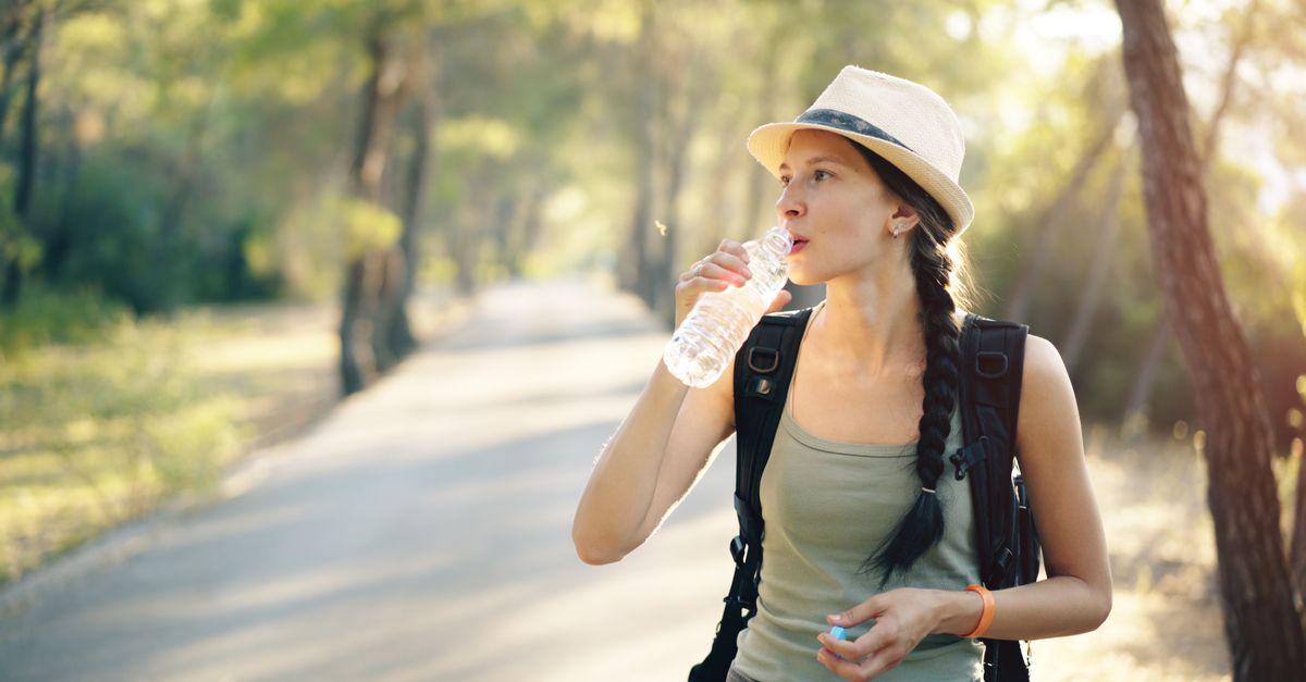 Young woman hiker in tank top and hat drinking water on a forest trail.