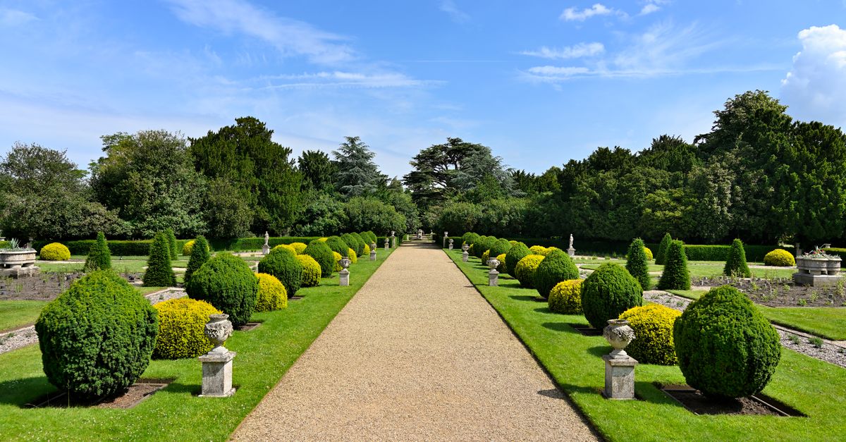 Elegant garden pathway lined with manicured bushes and classic pedestals under a clear sky.