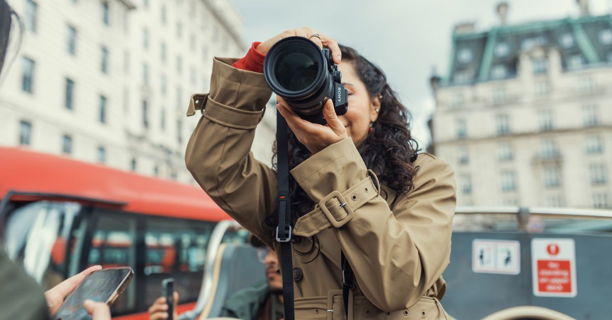 A woman with a camera takes photos from an open-top city tour bus in an urban setting.