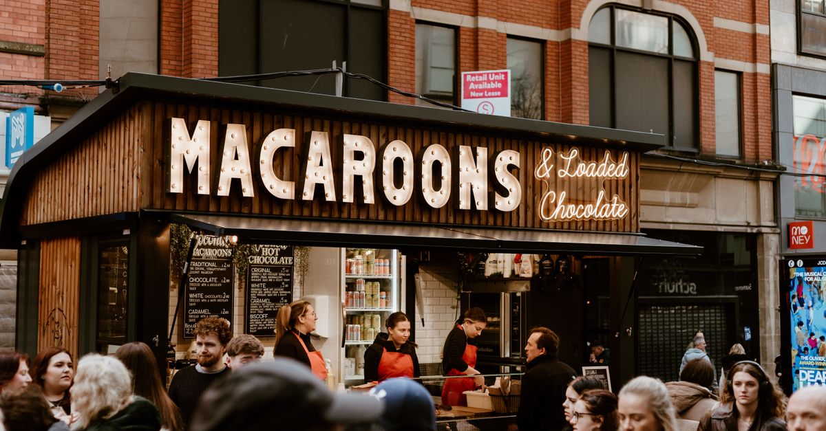 A bustling crowd gathers at an urban food stall selling macaroons and hot chocolate.