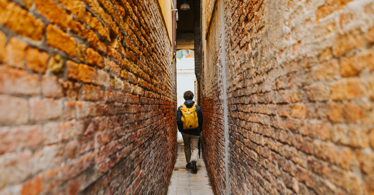A person with a yellow backpack walks through a narrow alley between brick walls, capturing a sense of exploration.