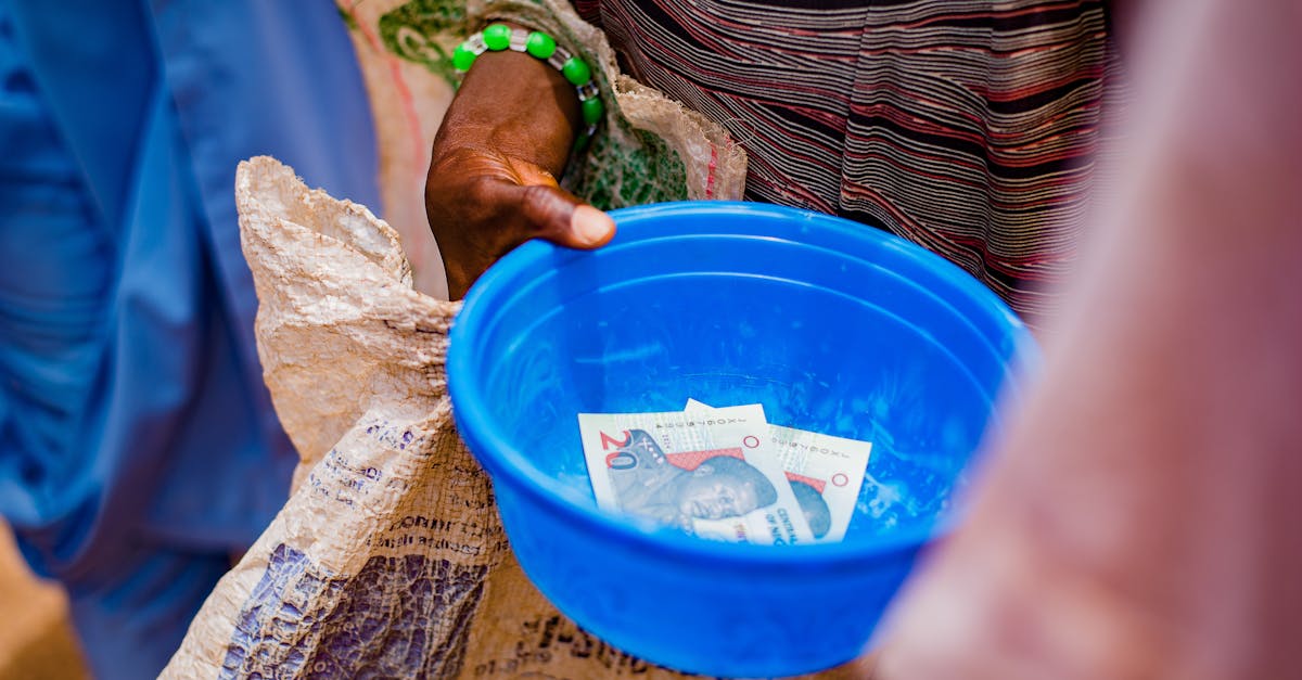 A close-up of a hand holding a blue bowl containing currency notes outdoors.