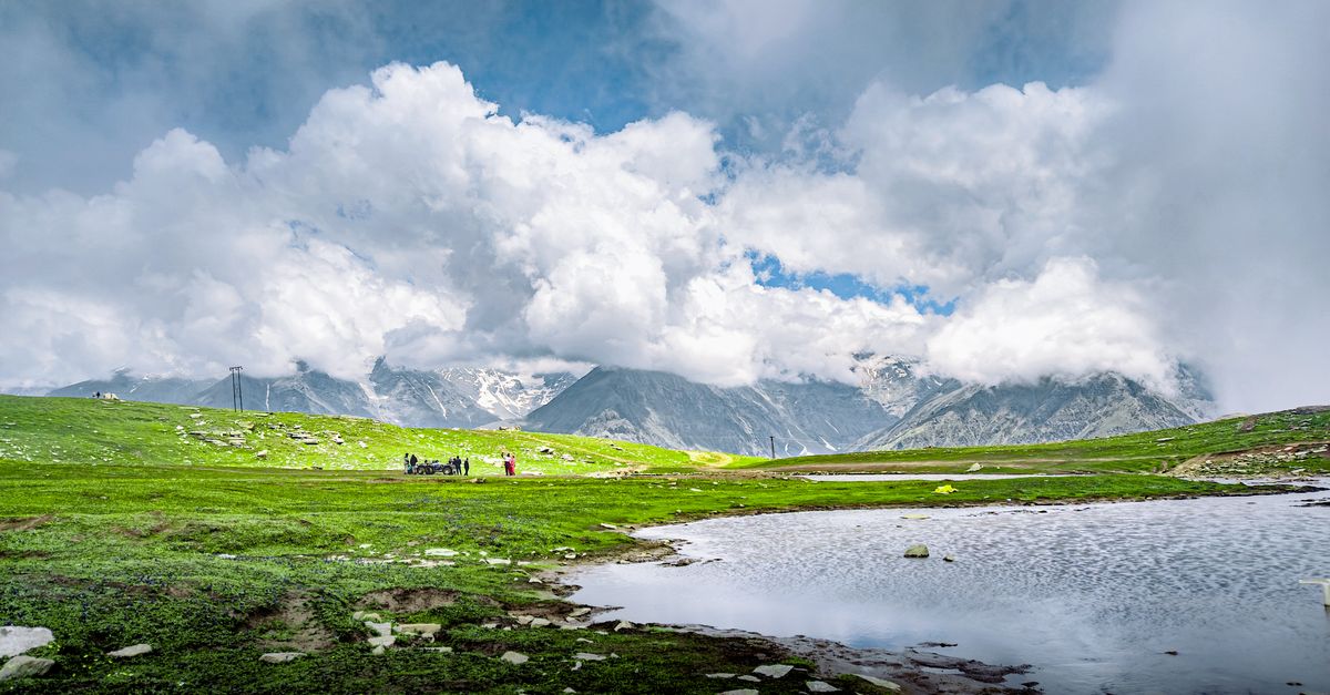 A breathtaking view of the Himalayas with lush green fields, a pristine lake, and dramatic clouds overhead.