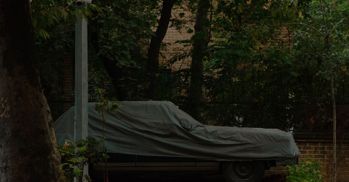 A vintage car covered with a tarp, parked outdoors against a brick wall, surrounded by trees.