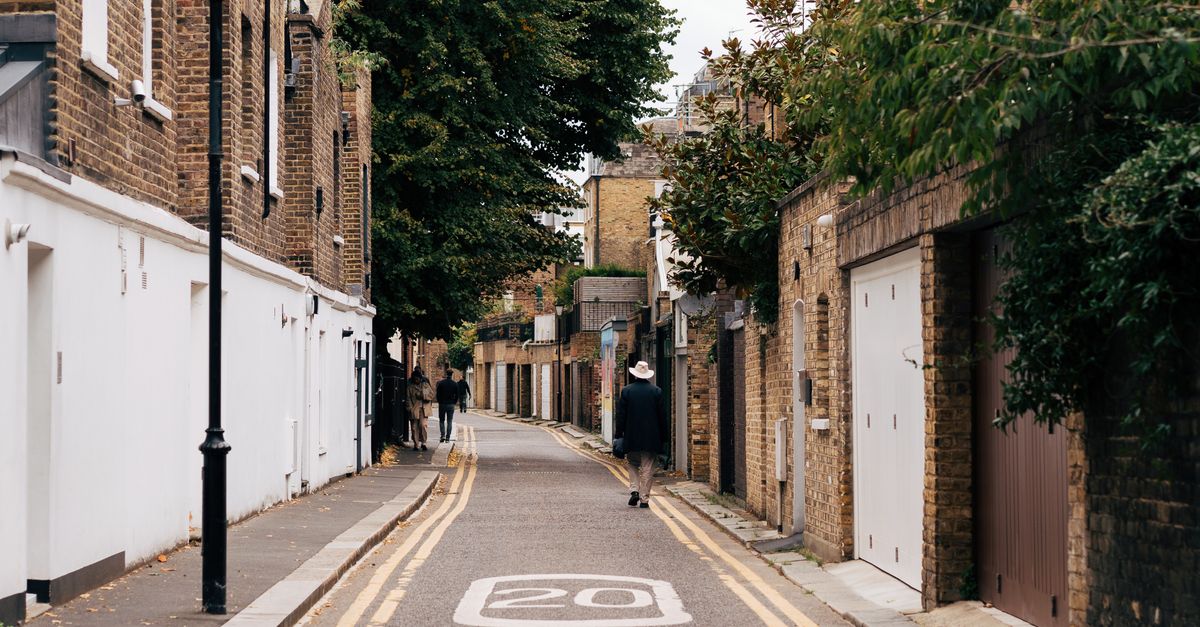 A quiet London street with brick houses, lush greenery, and a pedestrian walking.