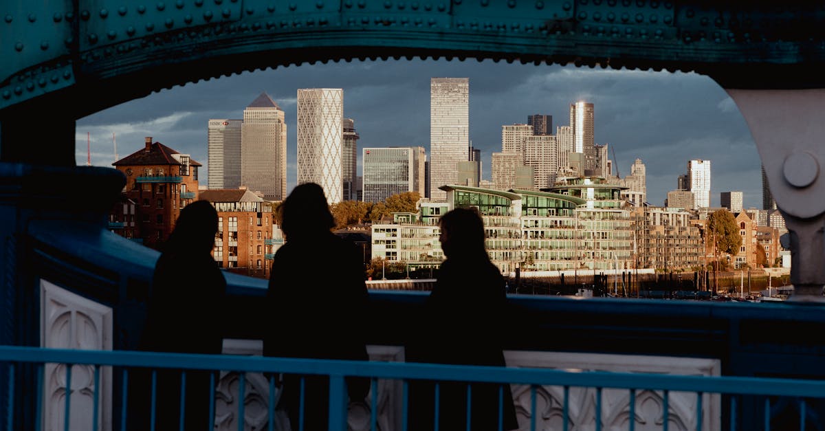 Three silhouetted figures frame a view of Canary Wharf's modern skyline at sunset from a London bridge.