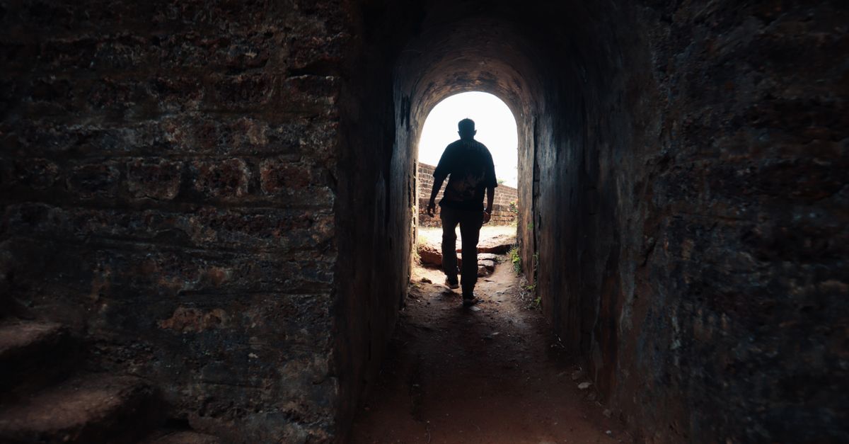 A solitary figure walks through a historic stone tunnel towards the light.