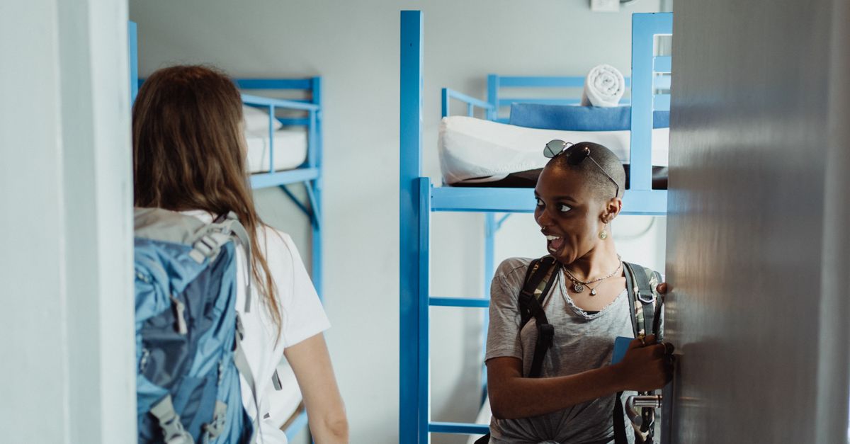 Two women with backpacks walking into a bright hostel dormitory with bunk beds, ready for adventure.