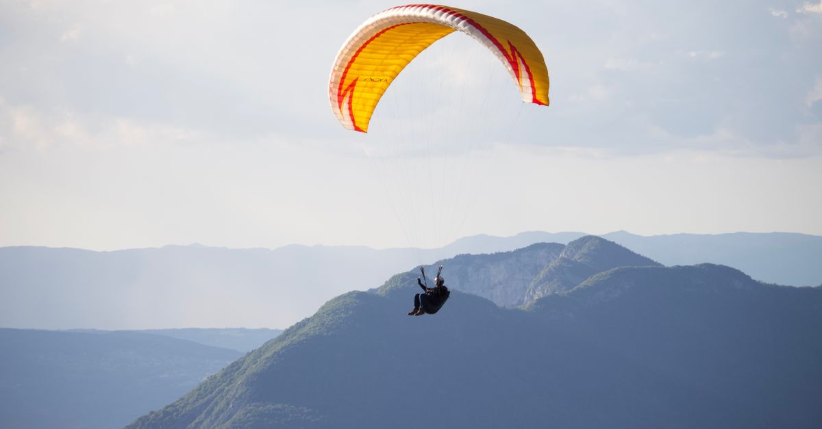 Unrecognizable distant person paraglider with yellow parachute flying high in air over mountainous area against cloudy sky during practicing in nature
