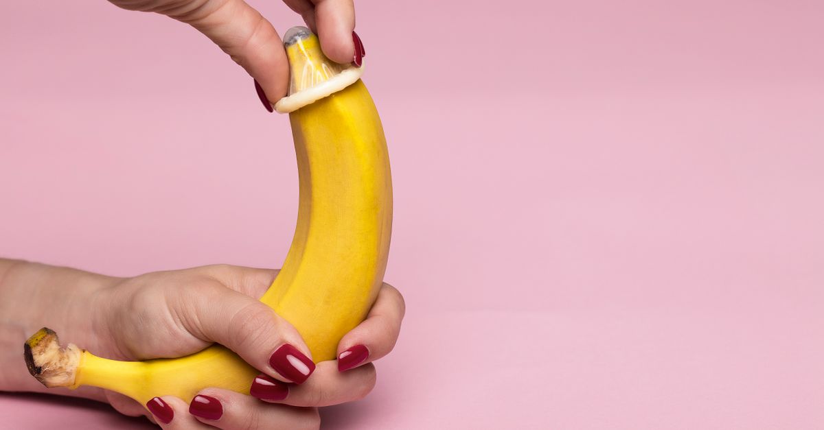 Close-up of hands applying a condom on a banana against a pink background symbolizing safe sex.