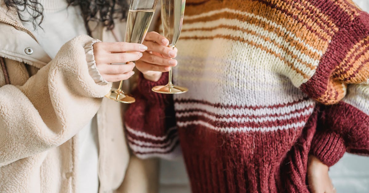 Crop faceless female friends in knitted sweaters clinking wineglasses of champagne during celebration together