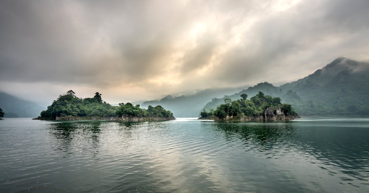 Picturesque scenery of rocky cliffs in rippling water of pond against green hills in mist under clouds