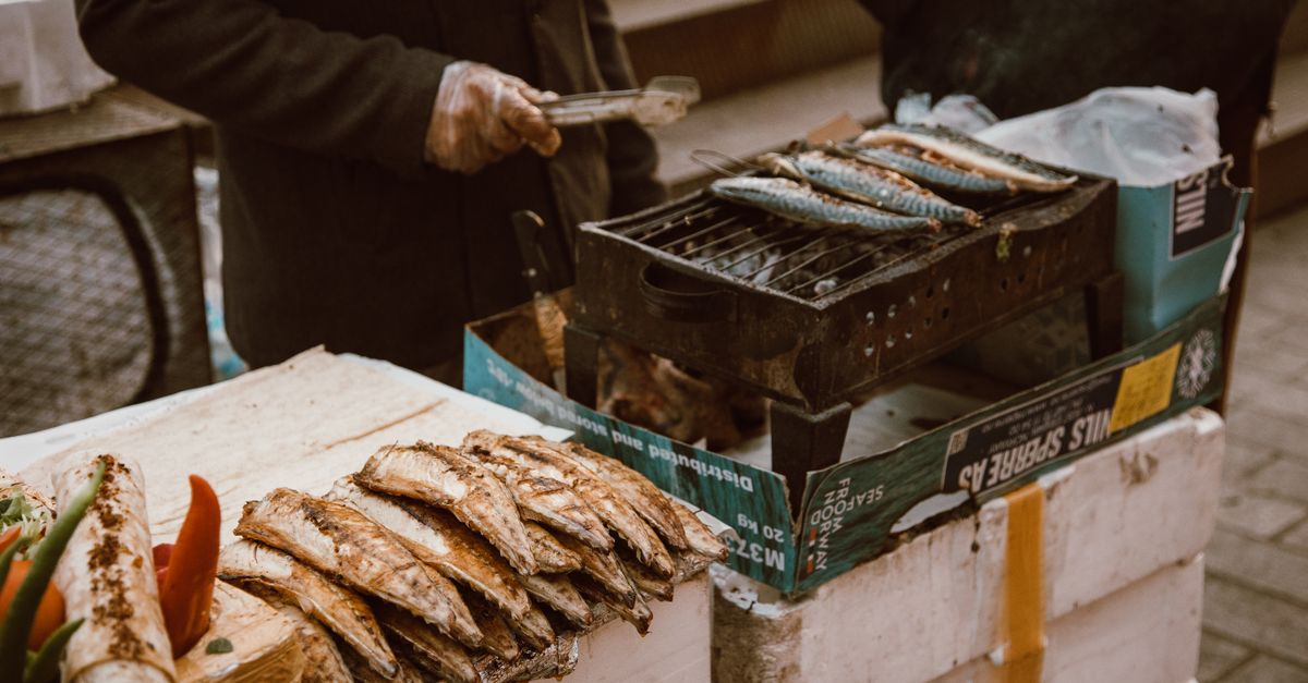 A street vendor grills fresh fish on a portable grill surrounded by fish and vegetables.