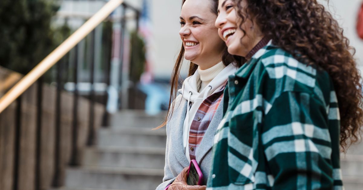 Side view of joyful young multiethnic female best friends with dark hair in stylish outfits smiling happily looking away while standing on city street near staircase in daytime